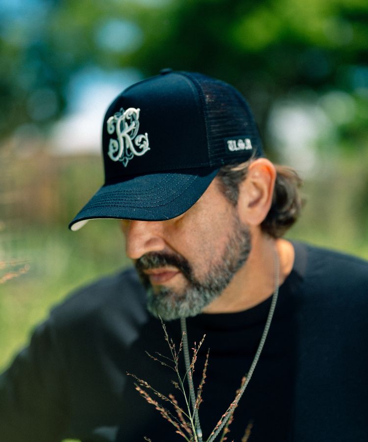 A man wearing a black trucker-style baseball cap with a white embroidered monogram logo on the front and 'USA' on the side by Philip Karto, photographed outdoors with a blurred green background.