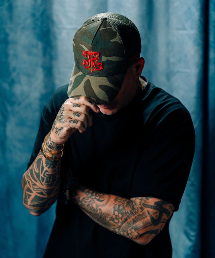 Person wearing a Camouflage PK Cap "Not For Everyone" Model by Philip karto with a red PK logo against a blue curtain background