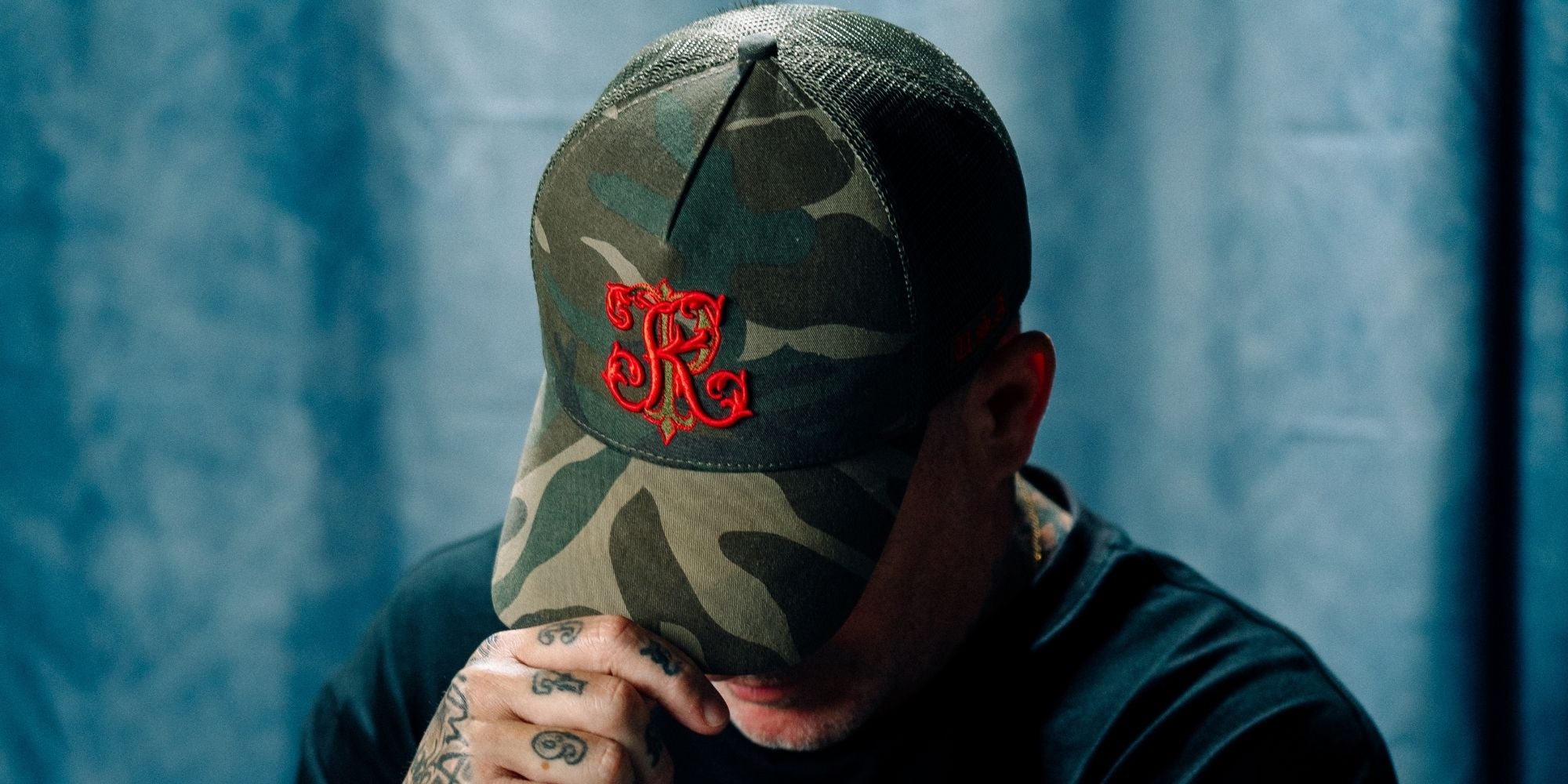 Philip Karto wearing a camouflage PK cap from the caps collection with a red PK logo against a blue background