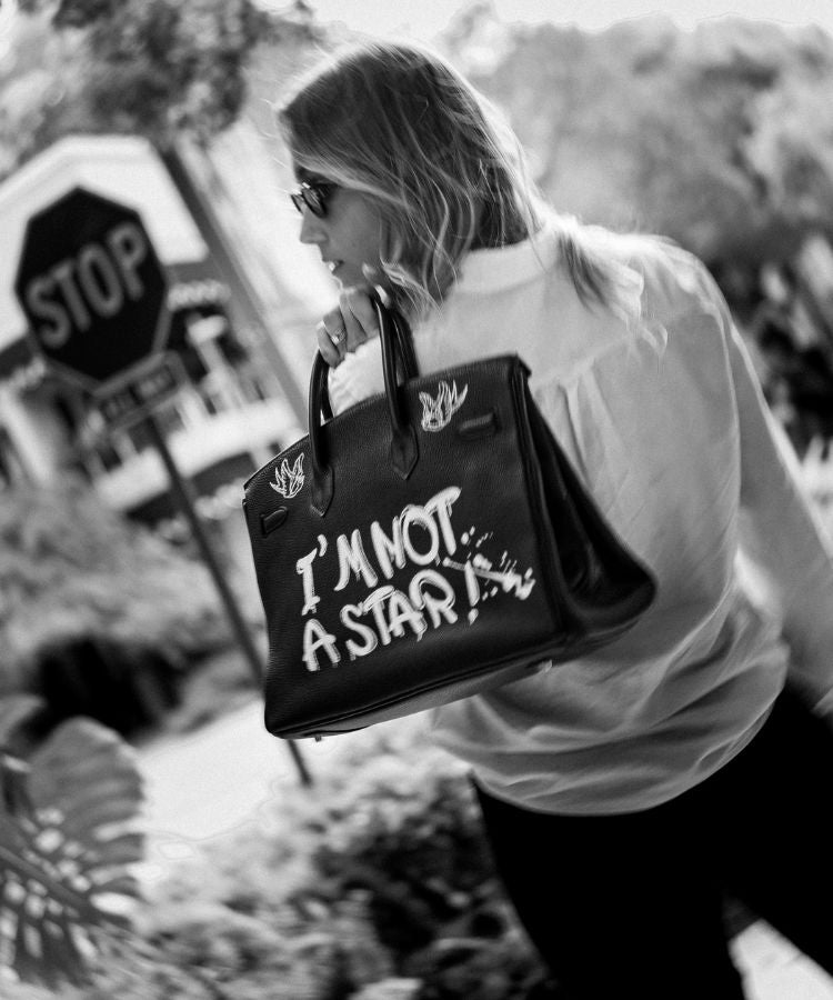 Person holding a black handbag w, outdoors. Person holding a large vintage Hermès handbag by Philip karto with 'I'm Not A Star' text