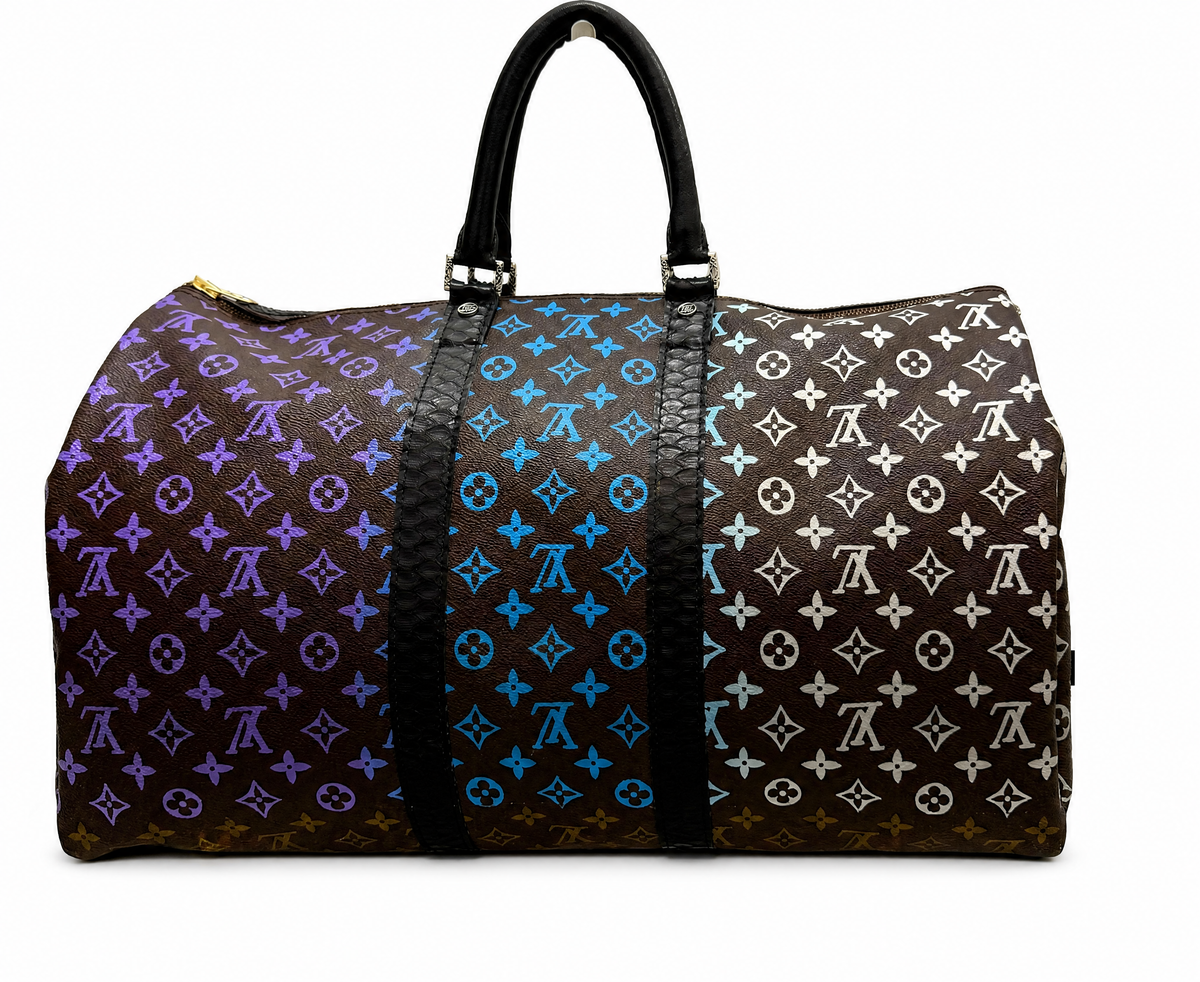 A Louis Vuitton monogram duffle bag with a tri-color gradient transition on the monogram canvas, featuring purple on one side, blue in the middle, and white on the other side. The bag has black leather handles and a black textured central strap, photographed against a solid white background.