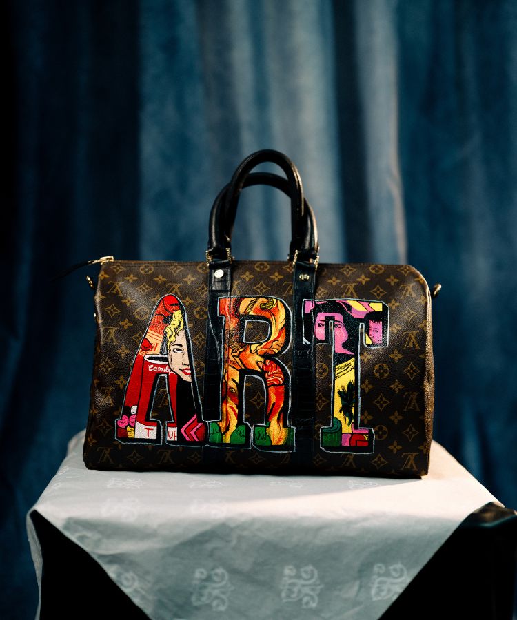 A Philip Karto monogram duffel bag with the word 'ART' spelled out in colorful patchwork letters, displayed on a draped white cloth against a dark blue curtain background.