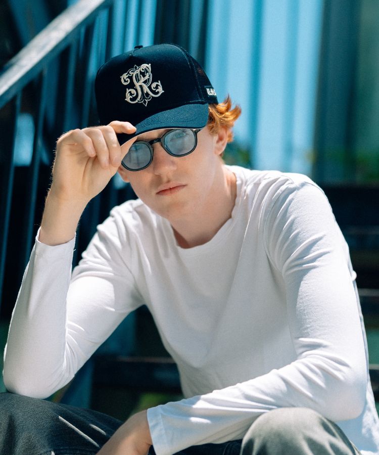 A young man wearing a black trucker cap with a white embroidered PK logo by Philip Karto, dark sunglasses, and a white long-sleeve shirt, sitting on outdoor steps.