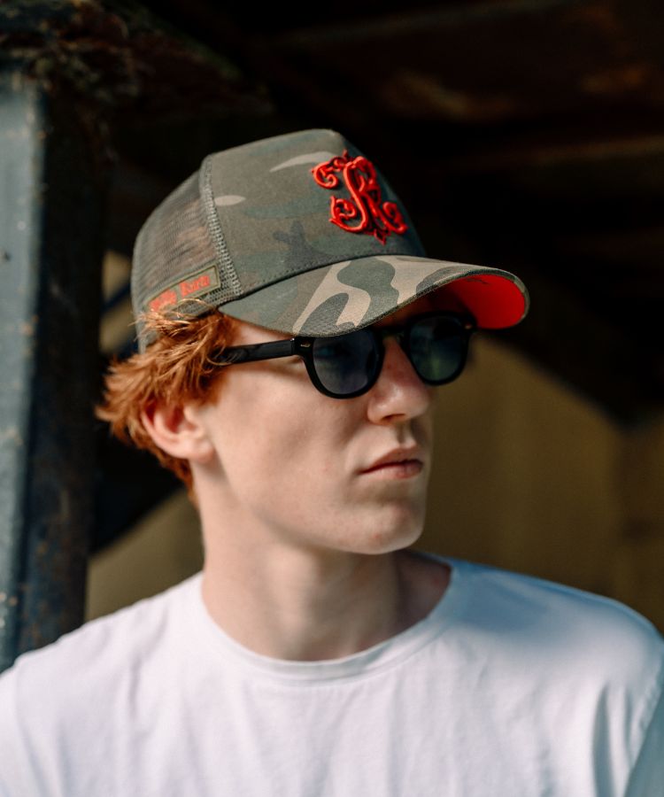 A young man with red hair wearing a green camouflage trucker hat with a red embroidered monogram logo on the front, black sunglasses, and a white t-shirt, captured in a warm, natural light setting.