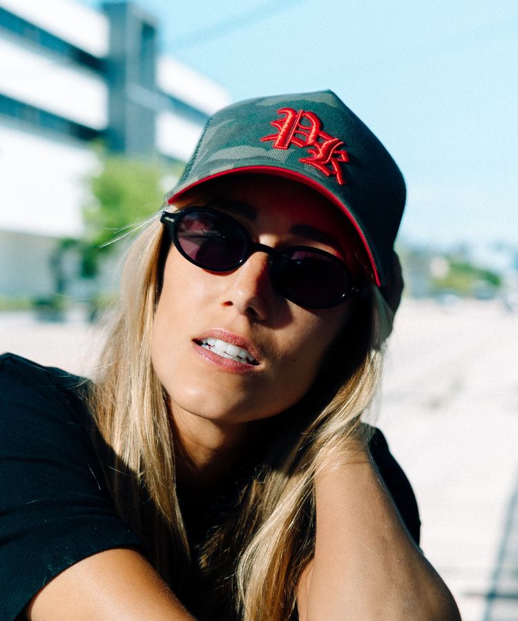 A woman wearing a Philip Karto Camouflage PK cap with red logo and sunglasses outdoors