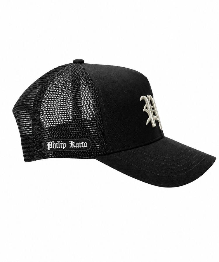 A black trucker-style baseball cap with a curved brim, black mesh back panels, a small white embroidered 'P' logo on the front, and a white 'Philip Karto' brand patch on the side.