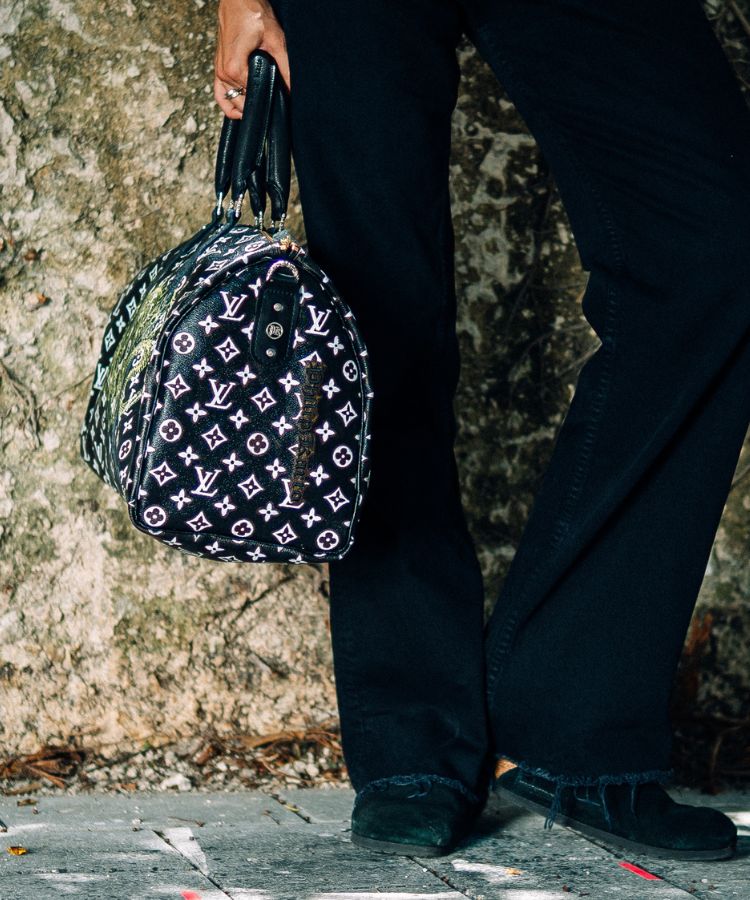 Woman holding a patterned Philip Karto handbag with Louis Vuitton monogram pattern and green snake design against a textured wall.