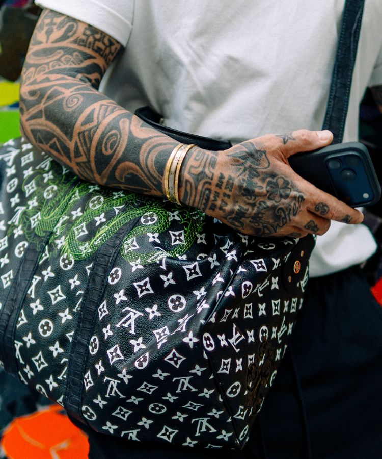 Person with tattooed arm holding a Philip Karto handbag with Louis Vuitton monogram pattern and green snake design
