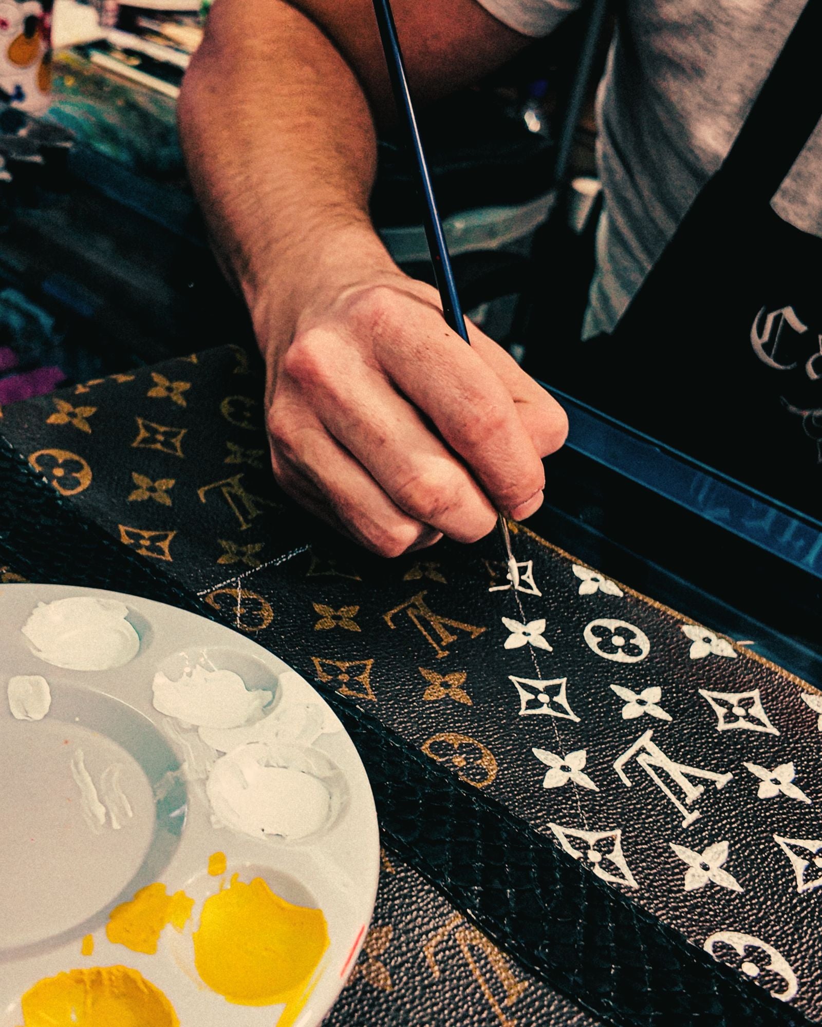 Hand of artist Philip Karto painting details over the monogram design on a Louis Vuitton canvas. It is a step of the bag creation with a paint palette and a brush.
